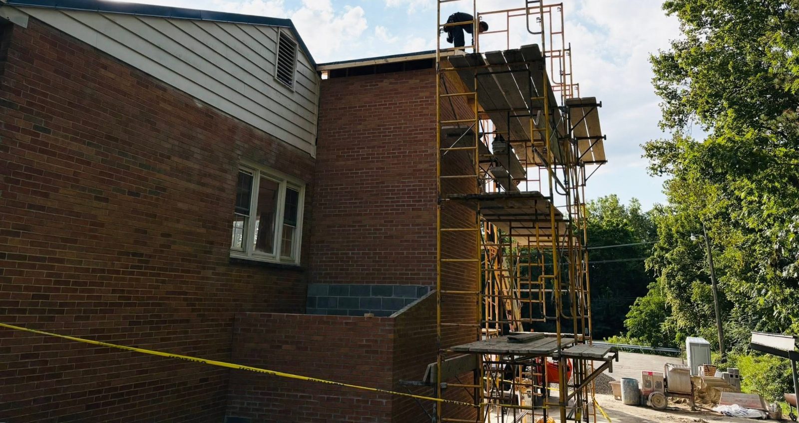 Exterior of American Legion Post 459 in Burton, Ohio during the accessibility project, with scaffolding along the brick wall and a worker on the roofline.